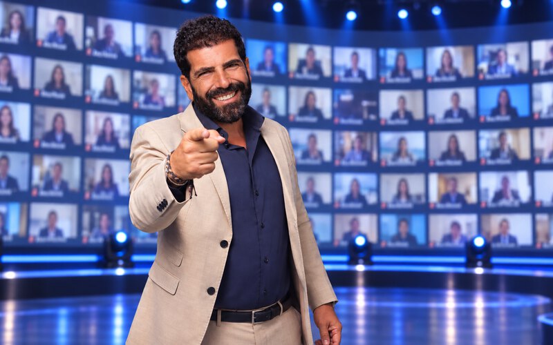 A host on stage in front of a wall of screens with the whole team joining online